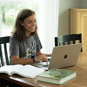 一个高中生坐在桌边微笑着用笔记本电脑工作